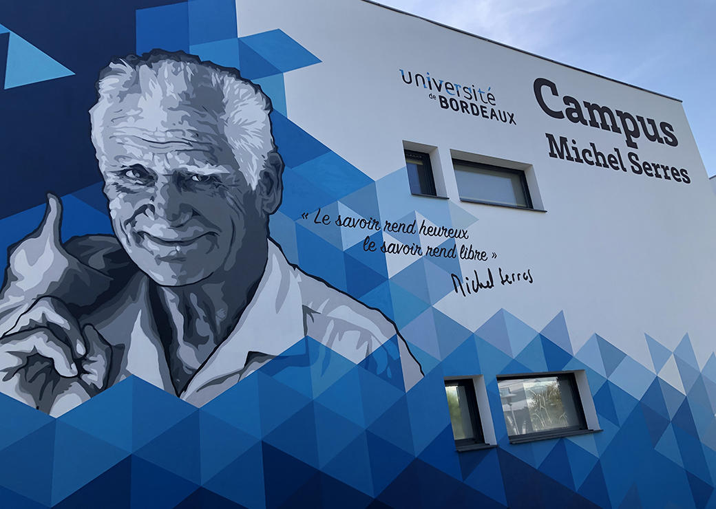 Cette image montre une fresque à base de bleus et de blanc représentant Michel Serres. Elle est affiché sur un bâtiment du campus Micel serres à Agen et contient une citation : "Le savoir rend libre, le savoir rend heureux".