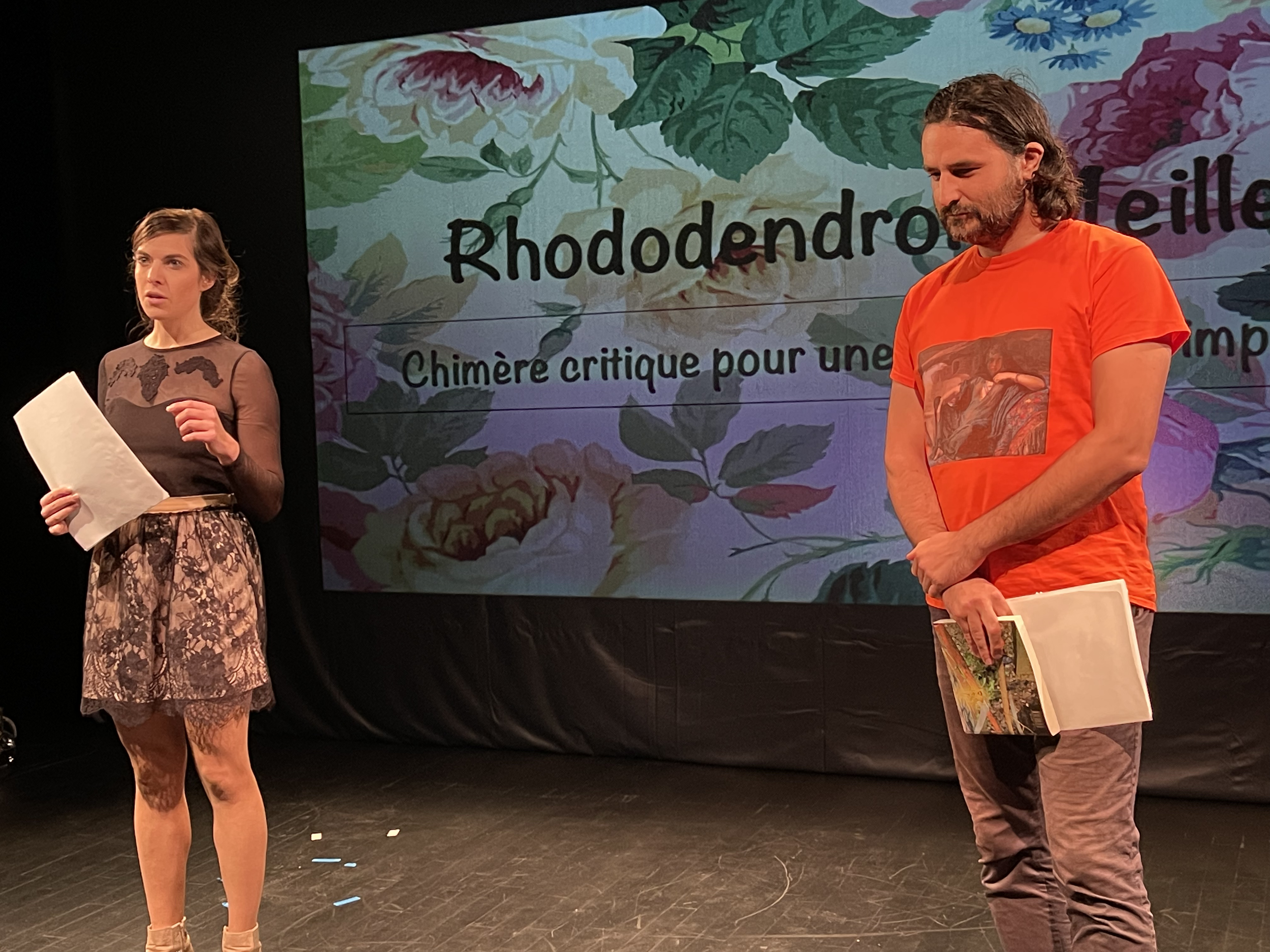 Cette photo montre deux artistes, un homme et une femme, debout devant un écran sur lequel est écrit Rhododendron. Ils tiennent tous les deux une feuille de papier