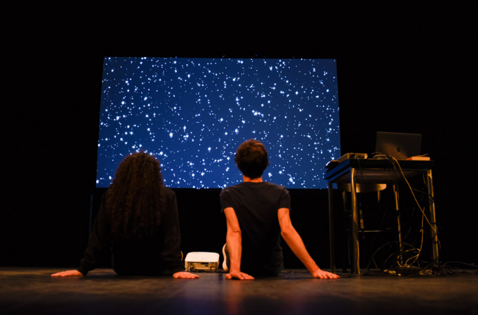 Cette photo montre un homme et une femme de dos assis sur une scène face à un écran affichant un ciel étoilé.