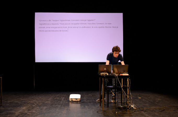 Cette Photo montre Simon Senn debout tapant sur le clavier d'un portable posé sur une table installée sur une scène. Derrière lui se trouve un écran sur lequel est projeté le contenu de l'écran du portable, un dialogue.